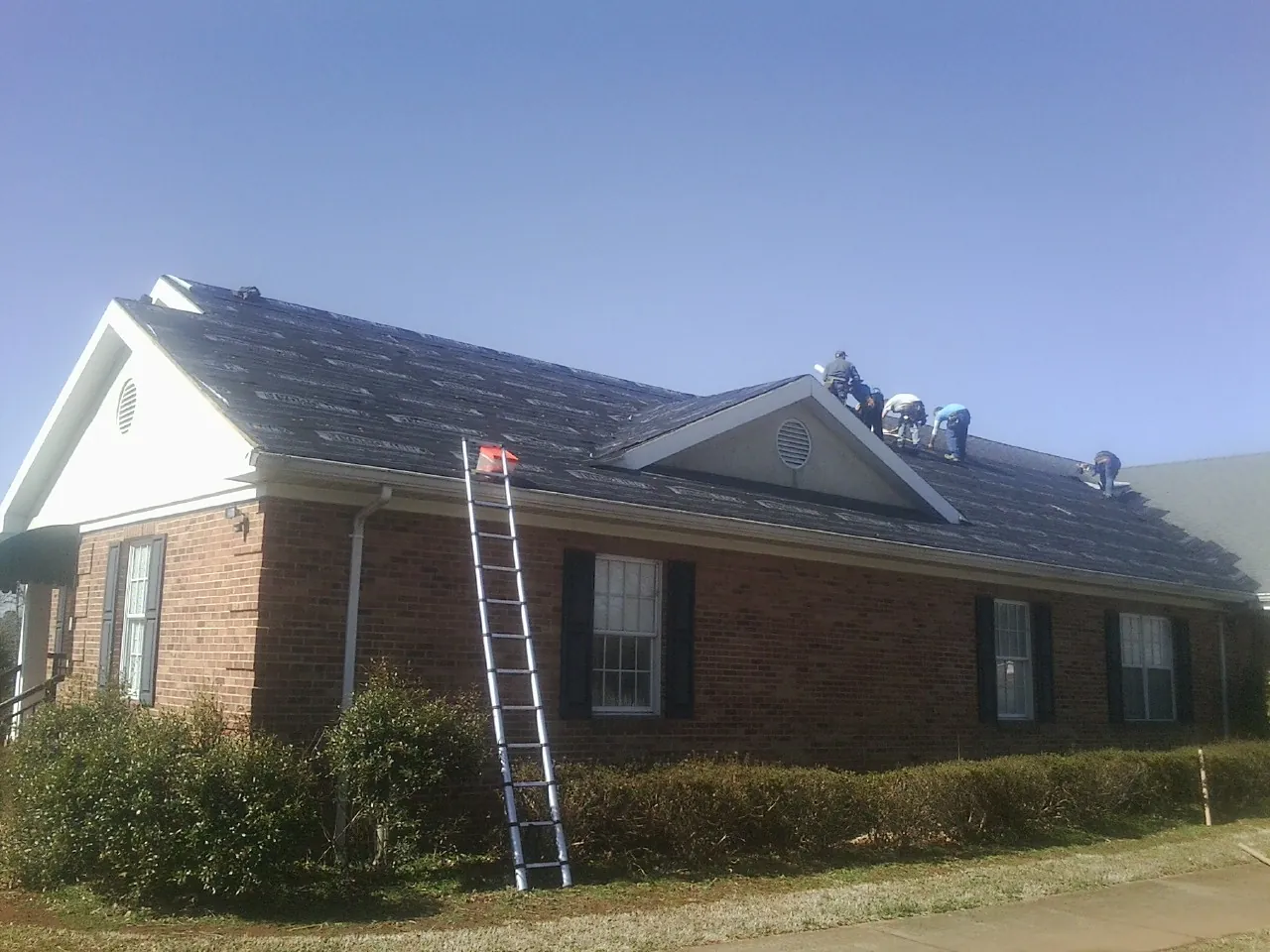 Crew tearing shingles off a brick ranch roof with a ladder set — Metal Roof Repair work by our crew in Penrose